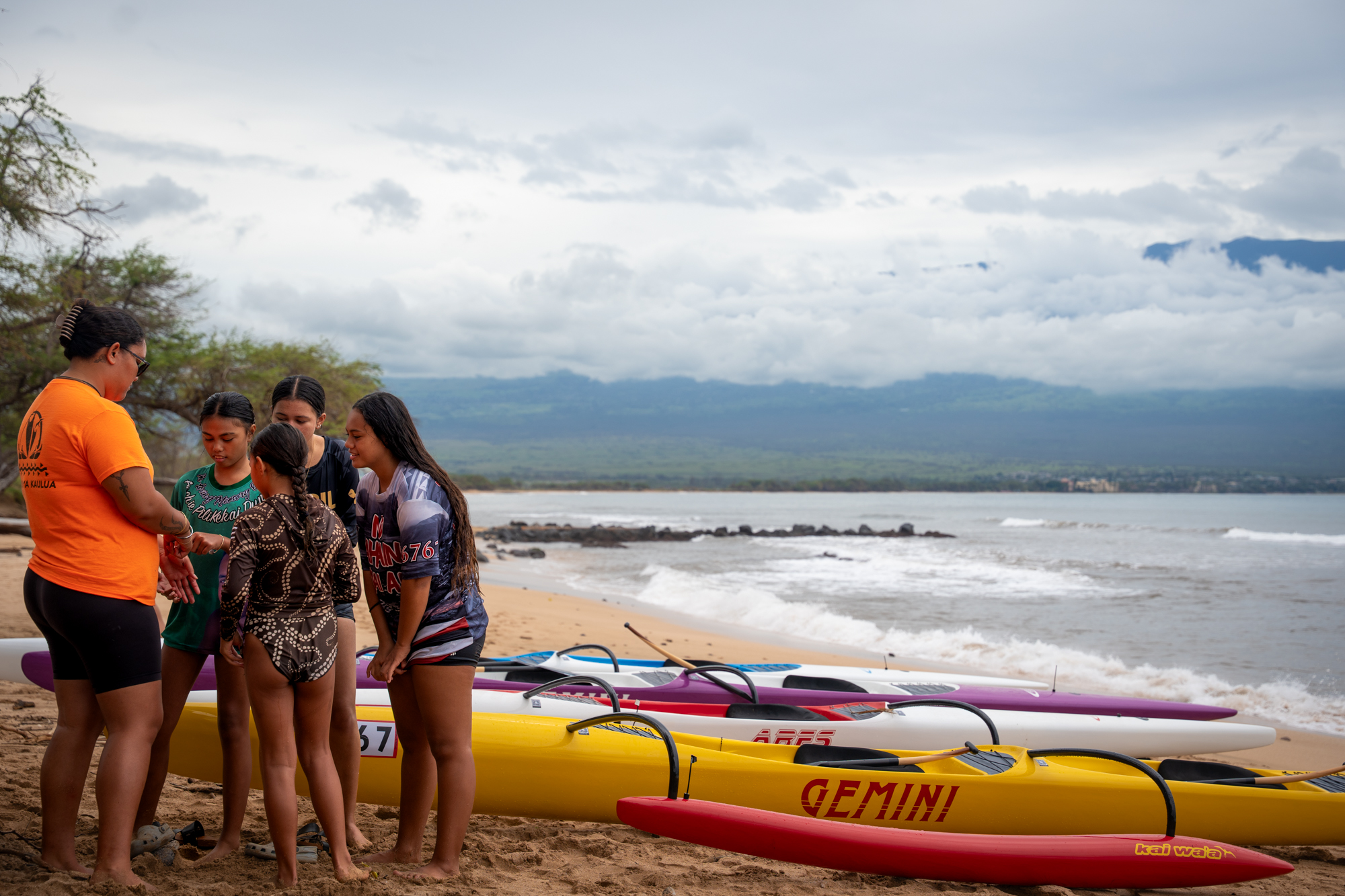 A young woman shows something to a group of young girls on a beach in front of three fiberglass canoes.