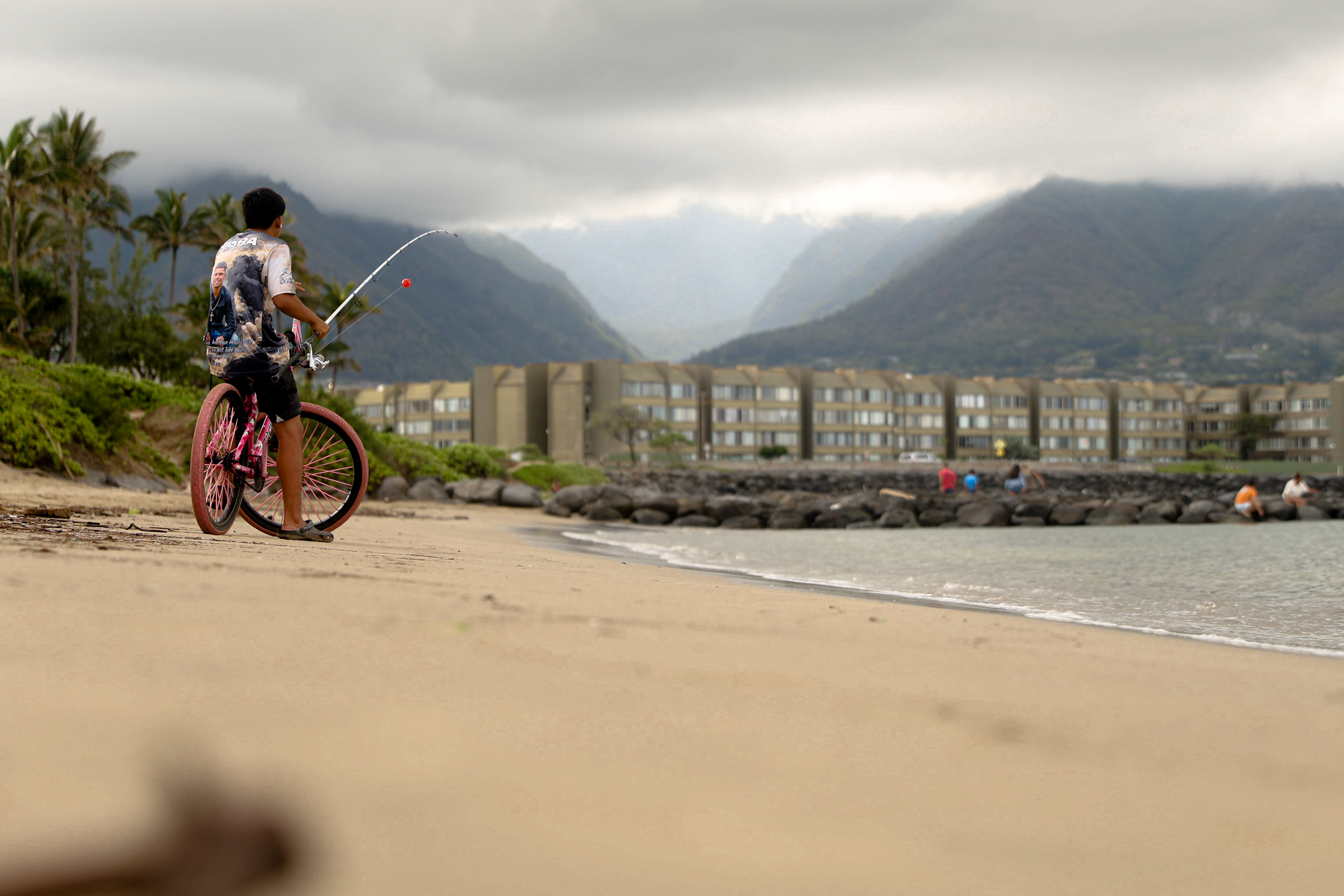 A young kid riding a bike on the beach with a fishing rod.