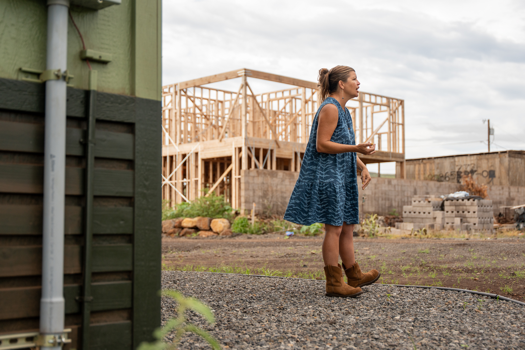 A woman in a blue dress and cowboy boots, Autumn Ness, stands in front of the wooden frame of a house under construction.