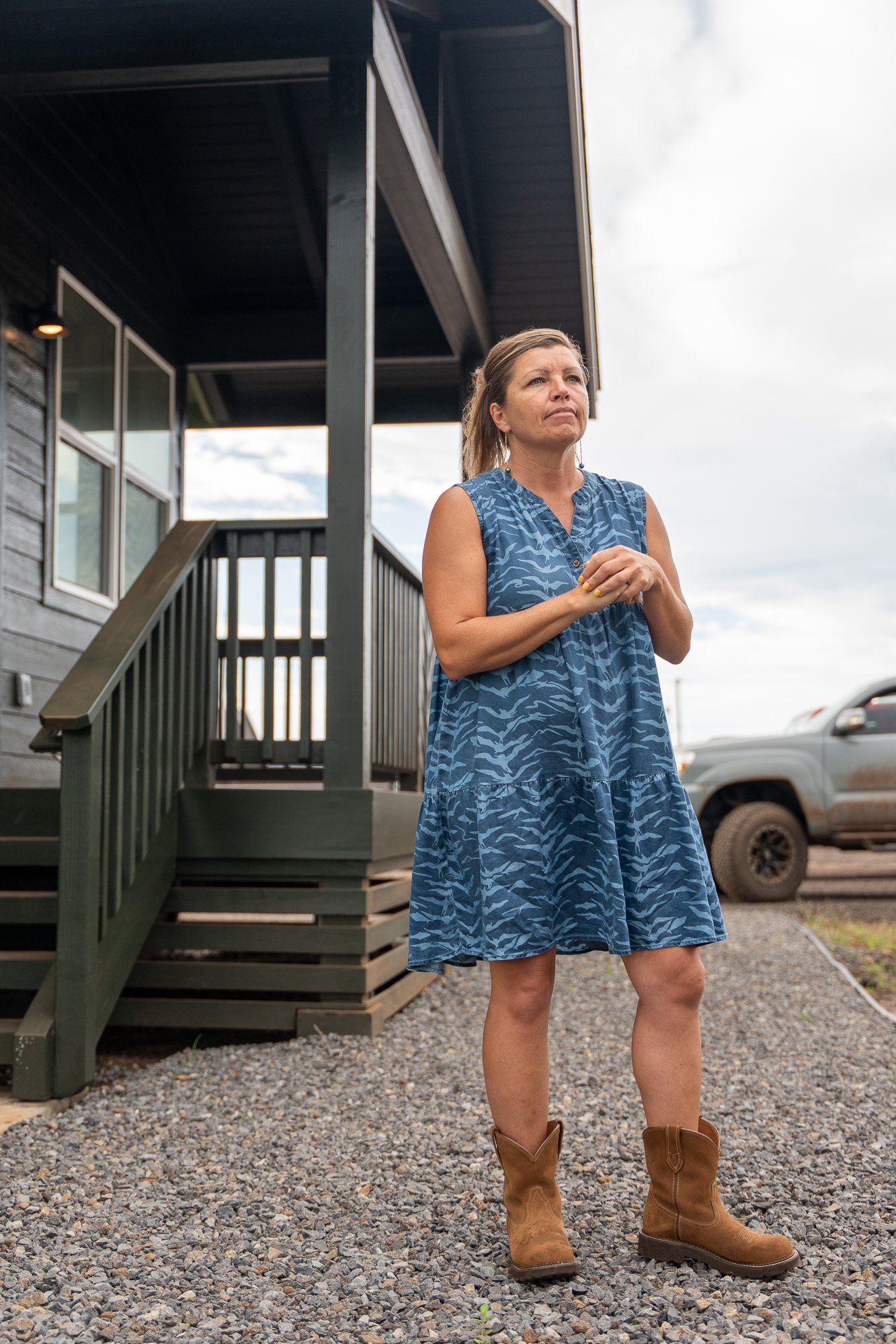 A woman in a blue dress and cowboy boots, Autumn Ness, stands in front of a house with a porch. Her mud-splattered truck in the background.